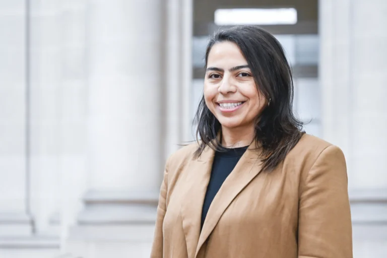 Smiling woman with dark hair in a tan blazer standing before large stone columns.