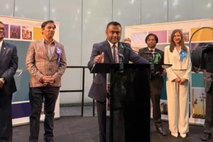 A man speaks at a podium with others wearing political rosettes standing behind him.