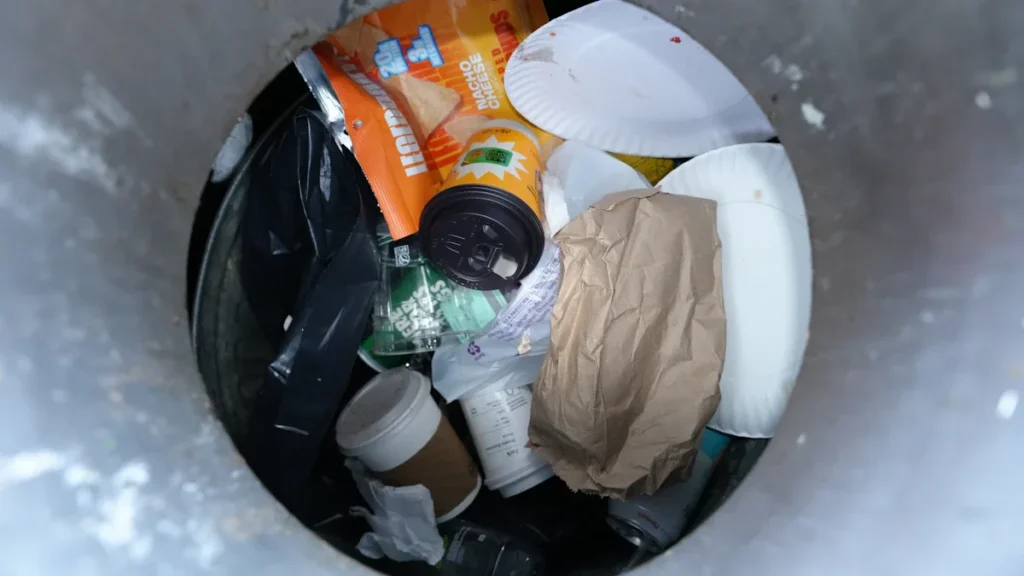 A top-down view into a waste bin filled with discarded paper plates, coffee cups, and food packaging, illustrating municipal waste.