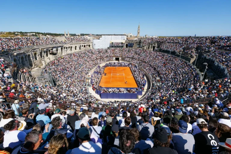 Wide view of a tennis match on a clay court inside a crowded, ancient stone amphitheater.