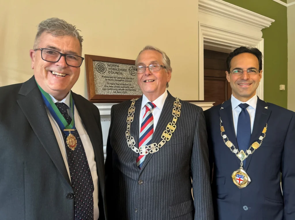 Three men wearing formal suits and ceremonial civic chains smile in front of a commemorative plaque.