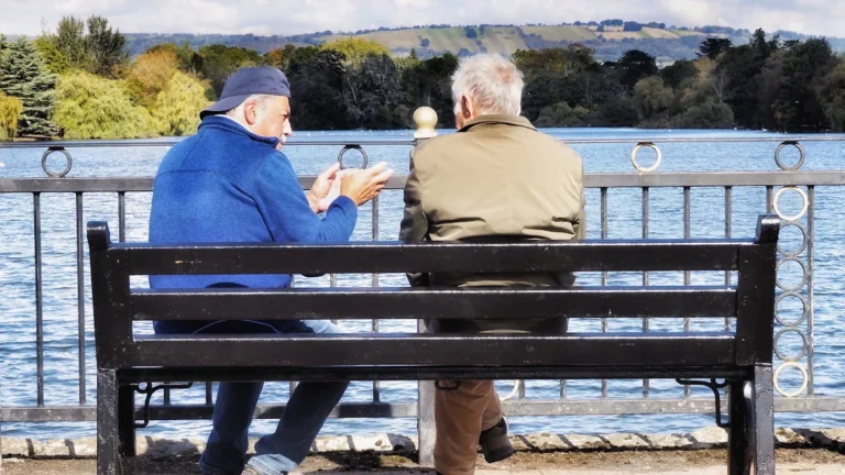 Two men sitting on a park bench by a lake, engaged in an earnest conversation.