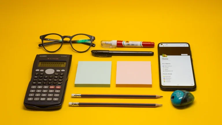 A smartphone sits alongside a calculator, pens, eyeglasses, and notebooks on a vibrant yellow background.