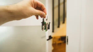 A hand holds house keys with a house-shaped keychain in a bright, modern residential hallway.