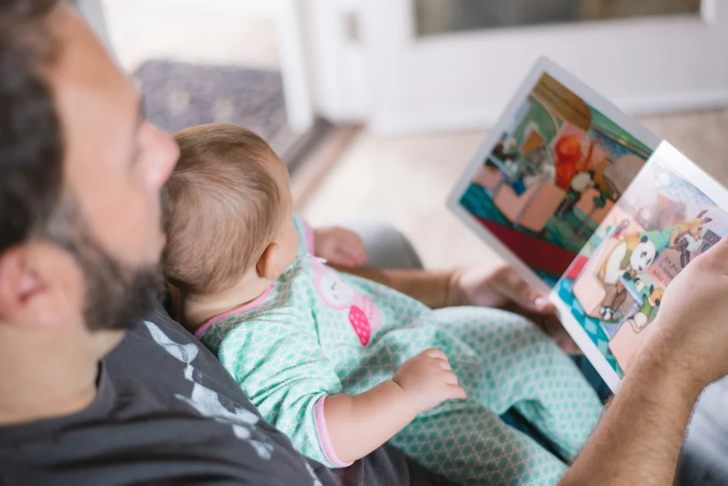 A man sits with an infant on his lap reading a colorful illustrated picture book.