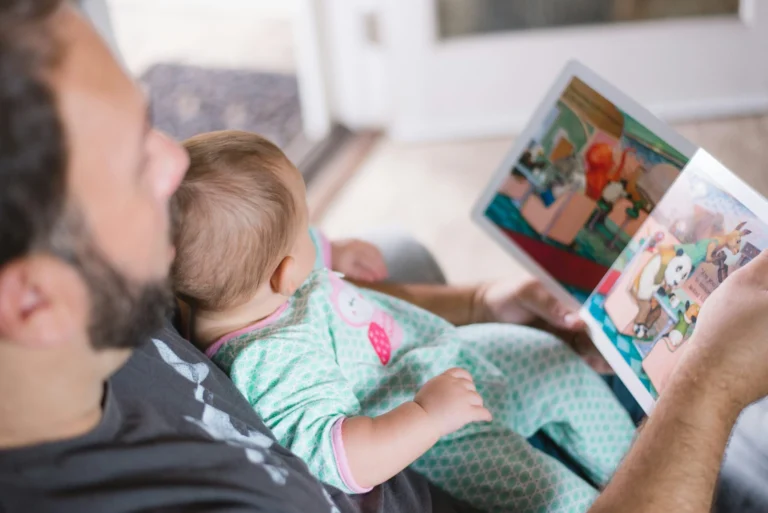 A man sits with an infant on his lap reading a colorful illustrated picture book.