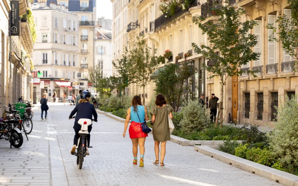 People walking and cycling on a sunny pedestrian street lined with historic stone buildings and greenery.