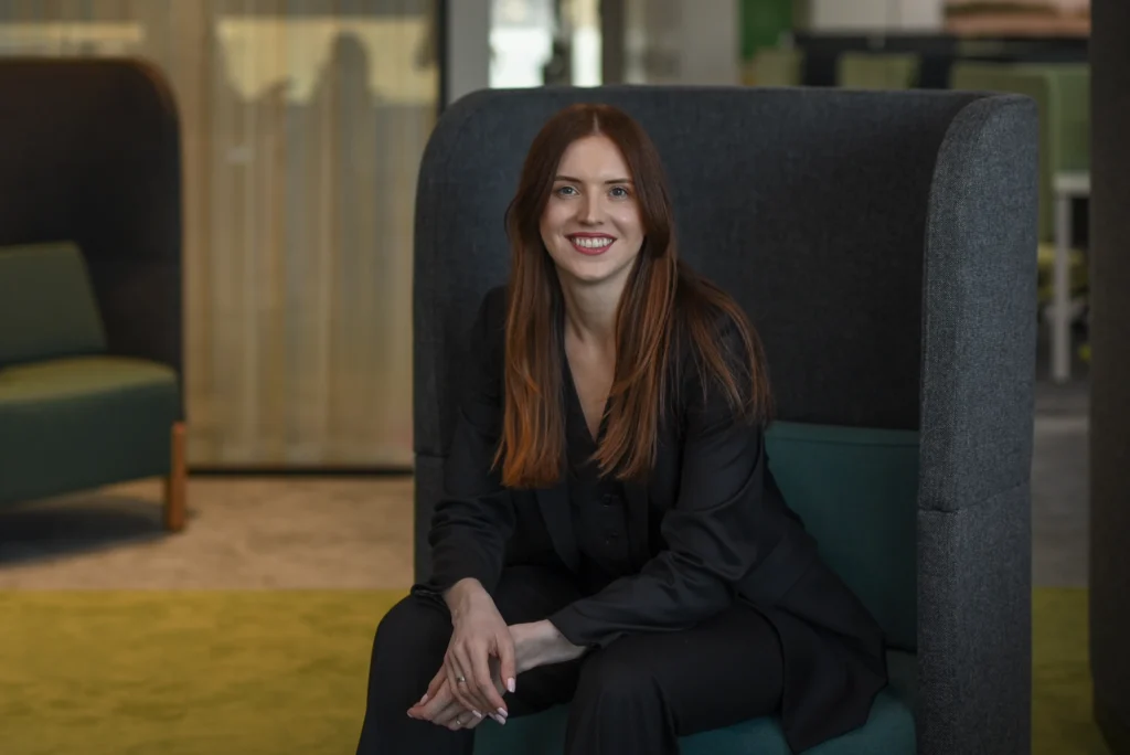 A professional woman with long reddish-brown hair smiles while sitting in a grey high-backed chair.