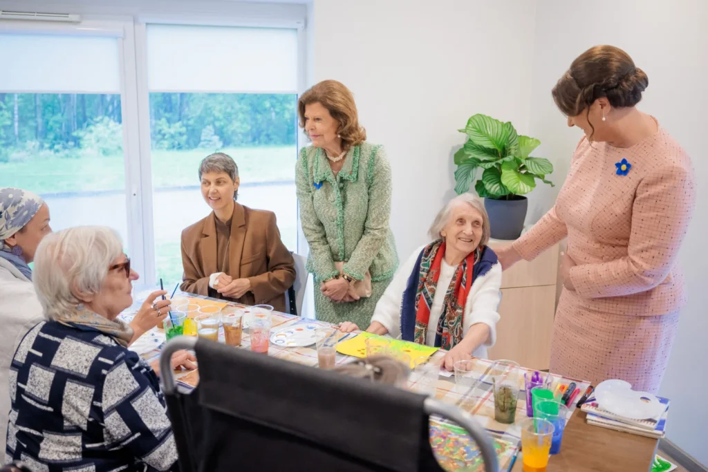 Queen Silvia and Agata Duda visit seniors engaged in a painting activity at a bright table.