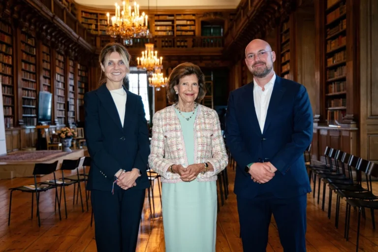 Three people standing together in a historic library with tall wooden bookshelves and ornate chandeliers.