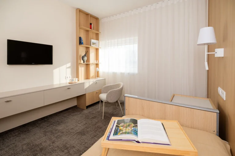 Minimalist room with a bed tray, television, wooden cabinetry, and light grey chair near a window.