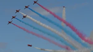 A formation of RAF Red Arrows jets flying across a blue sky with red, white, and blue smoke trails.