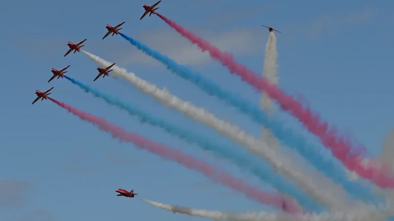 A formation of RAF Red Arrows jets flying across a blue sky with red, white, and blue smoke trails.