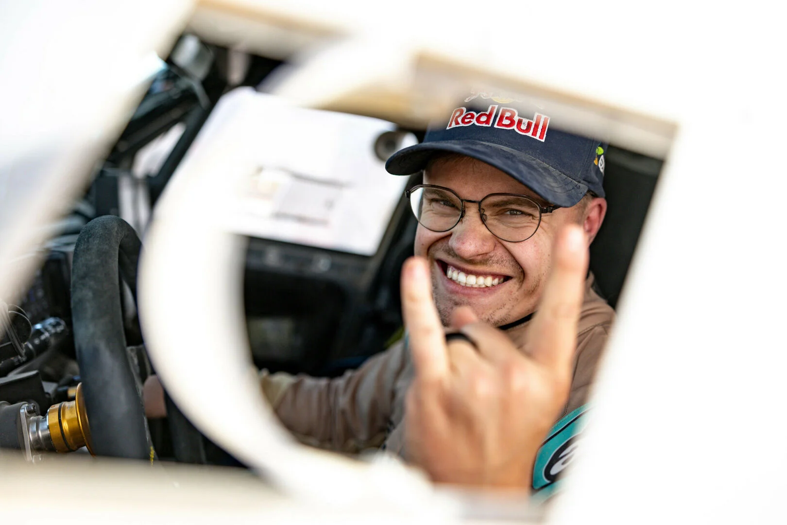 Smiling man in Red Bull cap making a rock hand gesture inside a racing car cockpit.