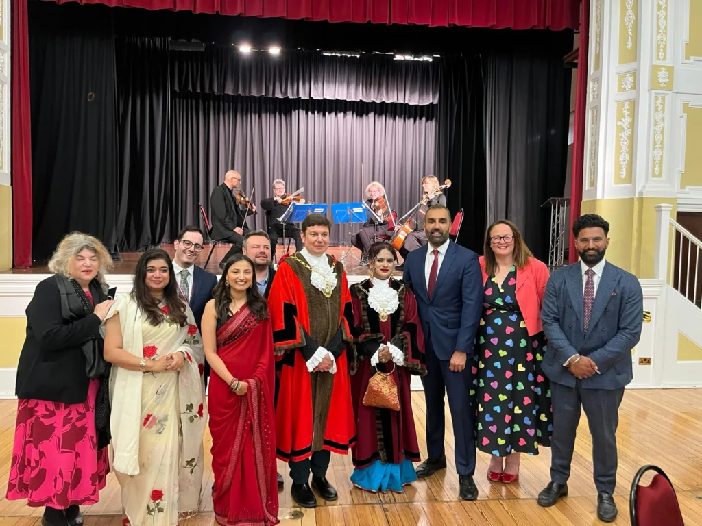 Diverse group in formal wear and mayoral robes posing before a live string quartet on stage.
