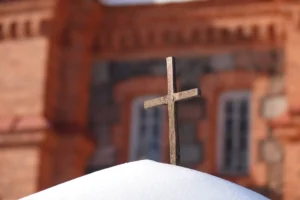 A weathered stone cross standing in white snow with a blurred red brick building background.