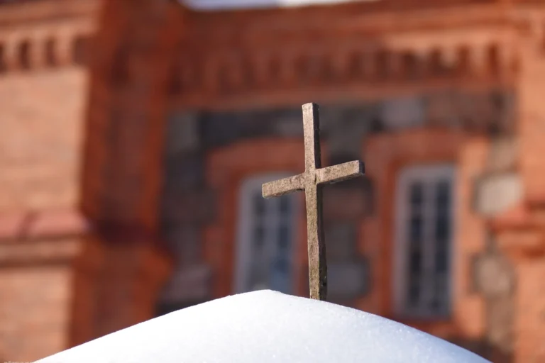 A weathered stone cross standing in white snow with a blurred red brick building background.