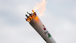 An Olympic-style torch with vibrant orange flames held diagonally against a pale, overcast sky.