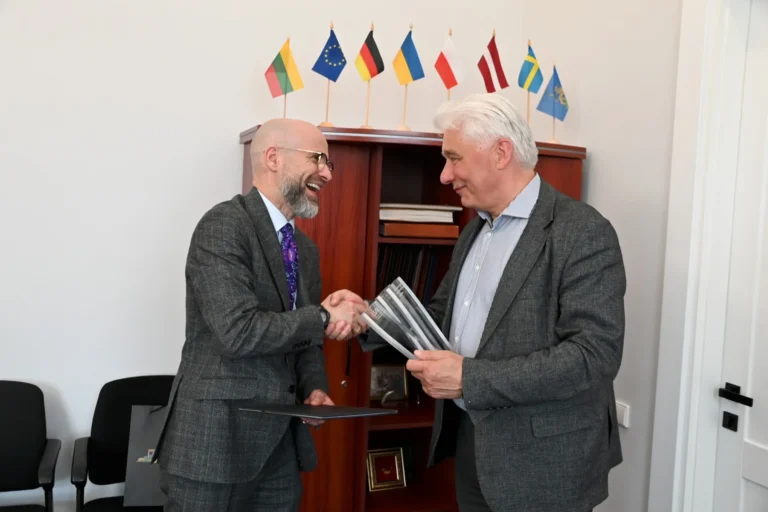 Two men in suits shake hands and smile in an office with international flags displayed.