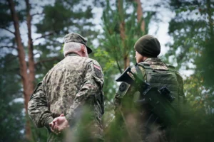 Two soldiers in camouflage uniforms, one older and one carrying a rifle, standing in a forest.
