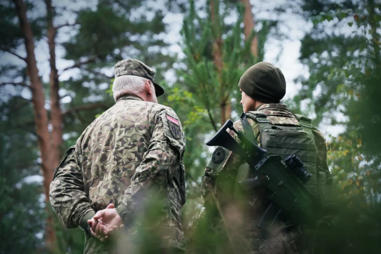 Two soldiers in camouflage uniforms, one older and one carrying a rifle, standing in a forest.