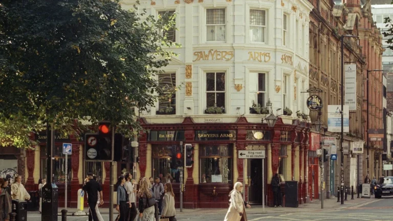 Pedestrians cross a busy Manchester street corner near a historic red-fronted pub during the day.