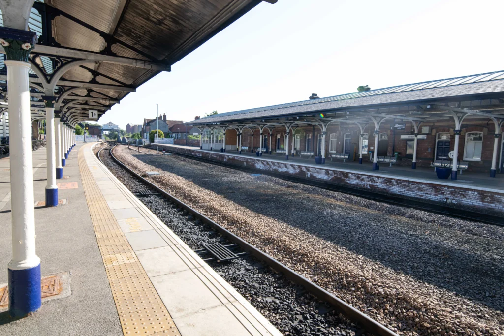 Empty train tracks and platforms with white Victorian-style roof supports under a bright, sunny sky.