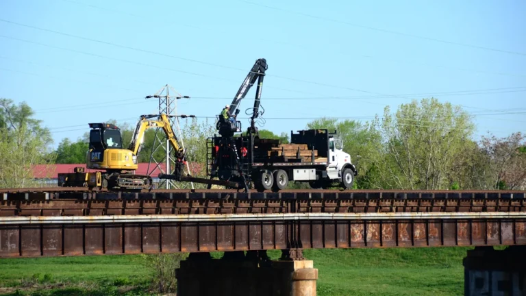 A construction crane and truck performing structural maintenance on a steel railway bridge.
