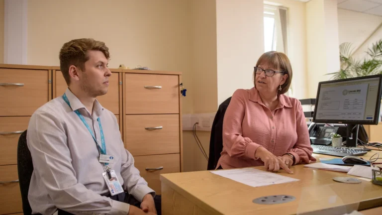 An older woman and a younger man discuss documents at a desk in a professional UK office.