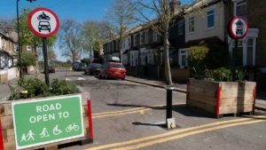 A UK residential street with a green sign reading Road Open To next to wooden planters.