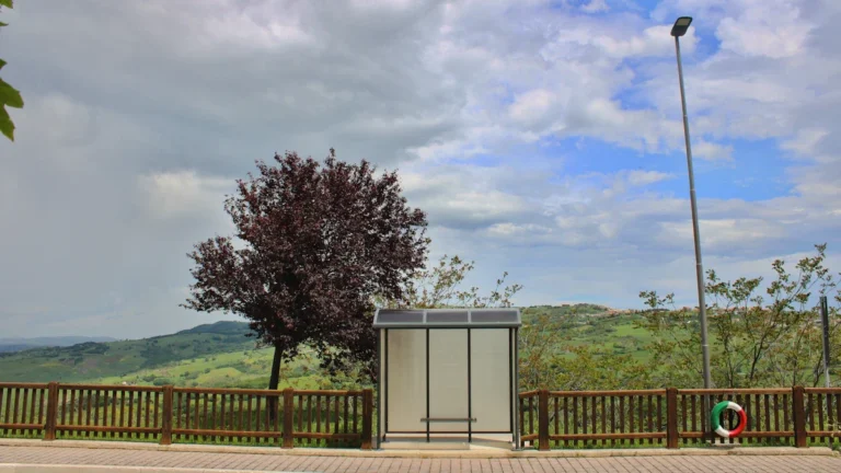 A lonely bus stop shelter positioned alongside a wooden fence overlooking a hilly rural landscape.