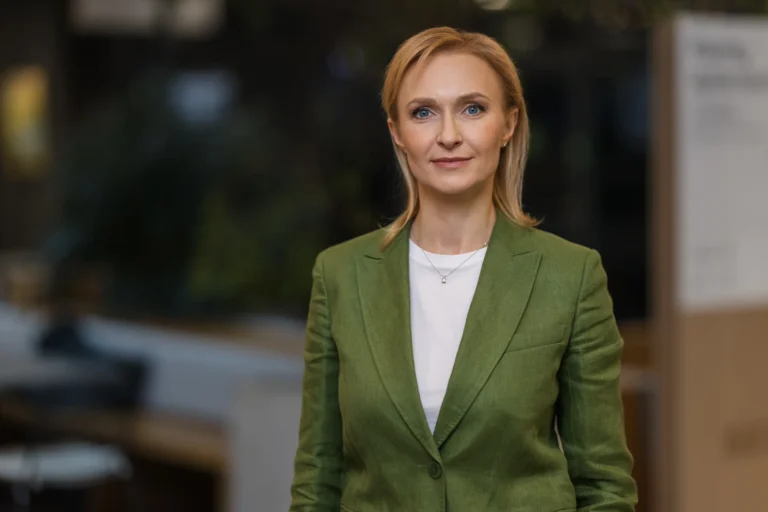 Blonde woman with blue eyes wearing a green blazer and white shirt in an office.