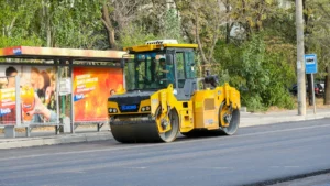 A yellow road roller smoothing fresh asphalt on a city street near a bus stop.