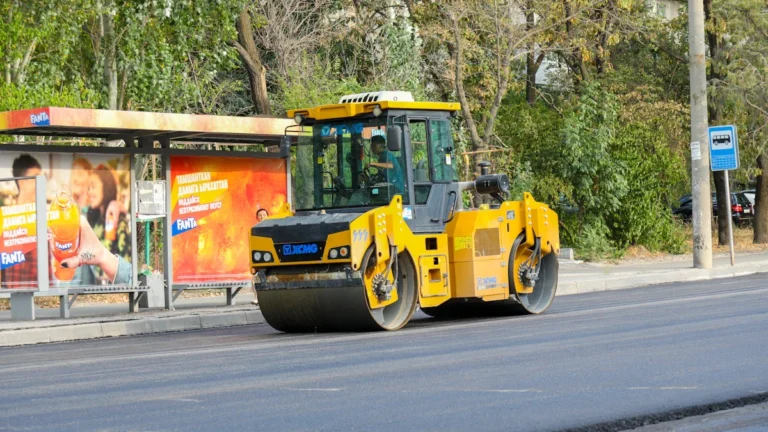 A yellow road roller smoothing fresh asphalt on a city street near a bus stop.
