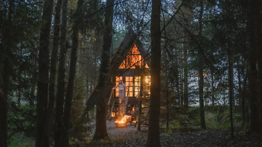 A small A-frame cabin glowing with warm light nestled deep within a dark pine forest.