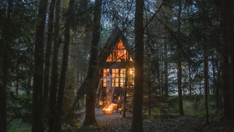 A small A-frame cabin glowing with warm light nestled deep within a dark pine forest.