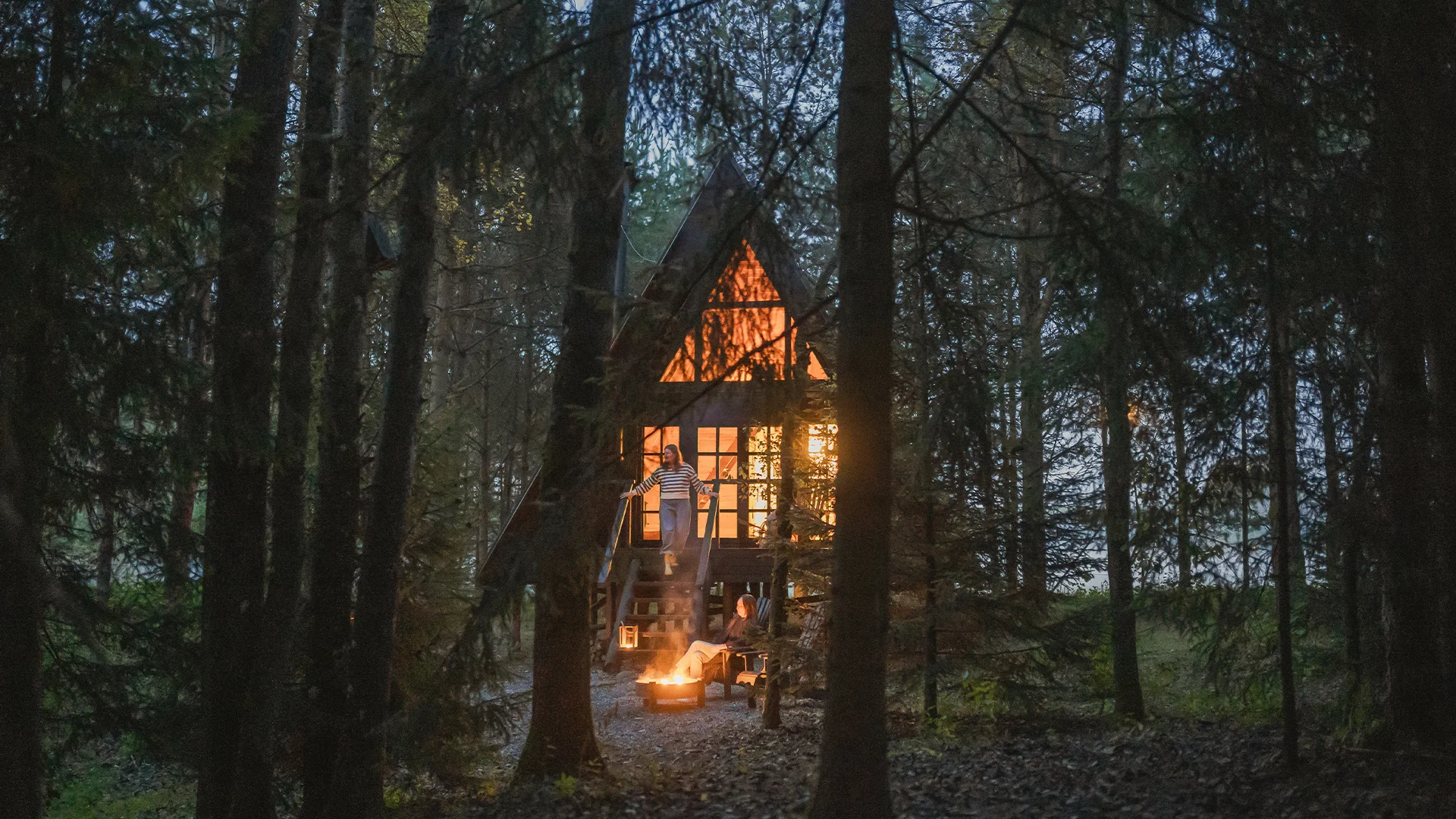 A small A-frame cabin glowing with warm light nestled deep within a dark pine forest.