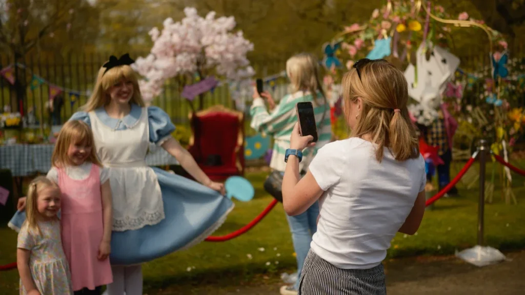 A woman takes a photo of two young children posing with a performer dressed as Alice in Wonderland at a sunny outdoor community event.