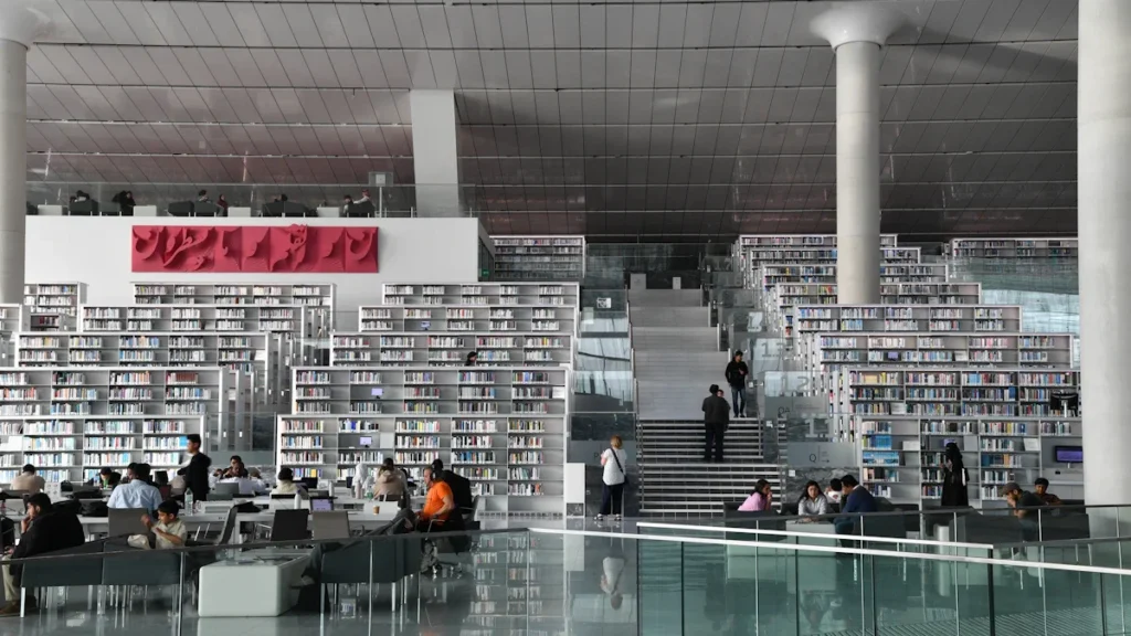 Modern library interior featuring tiered white bookshelves, large columns, and people in community seating areas.