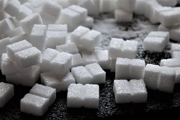 A pile of white refined sugar cubes scattered on a dark textured black background surface.