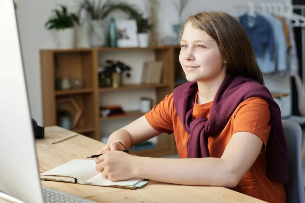 A teenage girl sitting at a desk looking at a computer screen with a notebook.