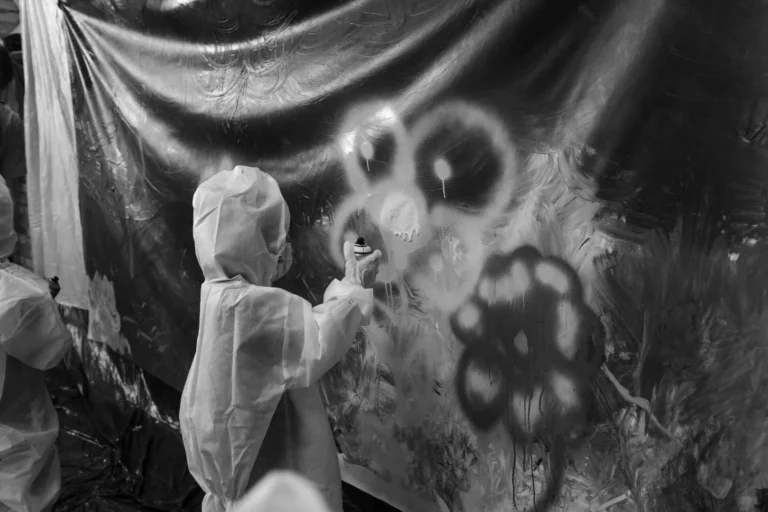 A child in white protective gear spray painting white flowers onto a large dark canvas.