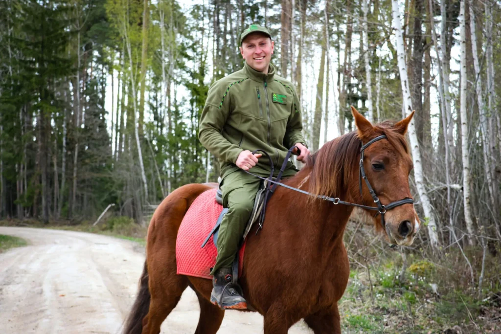 Smiling man in green forestry gear riding a brown horse on a dirt road through woods.