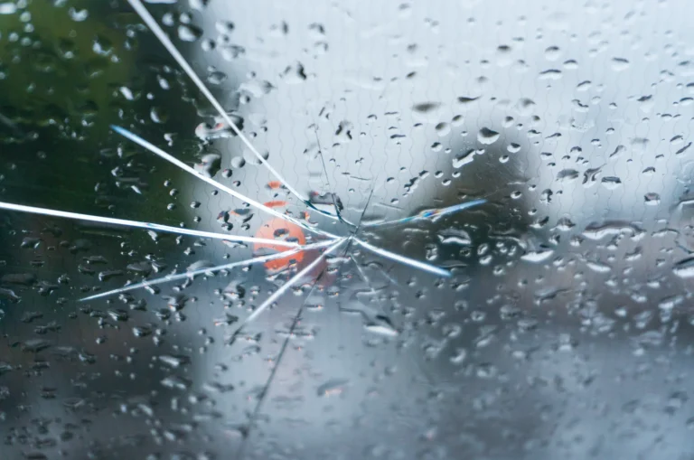 A cracked glass windshield covered in raindrops with several white cracks radiating from a central point.