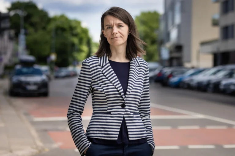 A woman with brown hair wearing a striped blazer stands outdoors on a city street.