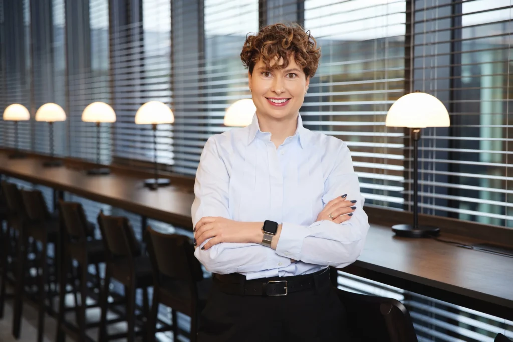 Smiling woman with curly brown hair standing with arms crossed in a modern office environment.