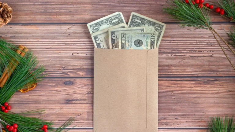 A top-down view of US dollar bills in a brown kraft envelope on a wooden table, framed by festive Christmas greenery and red berries, representing a holiday budget.