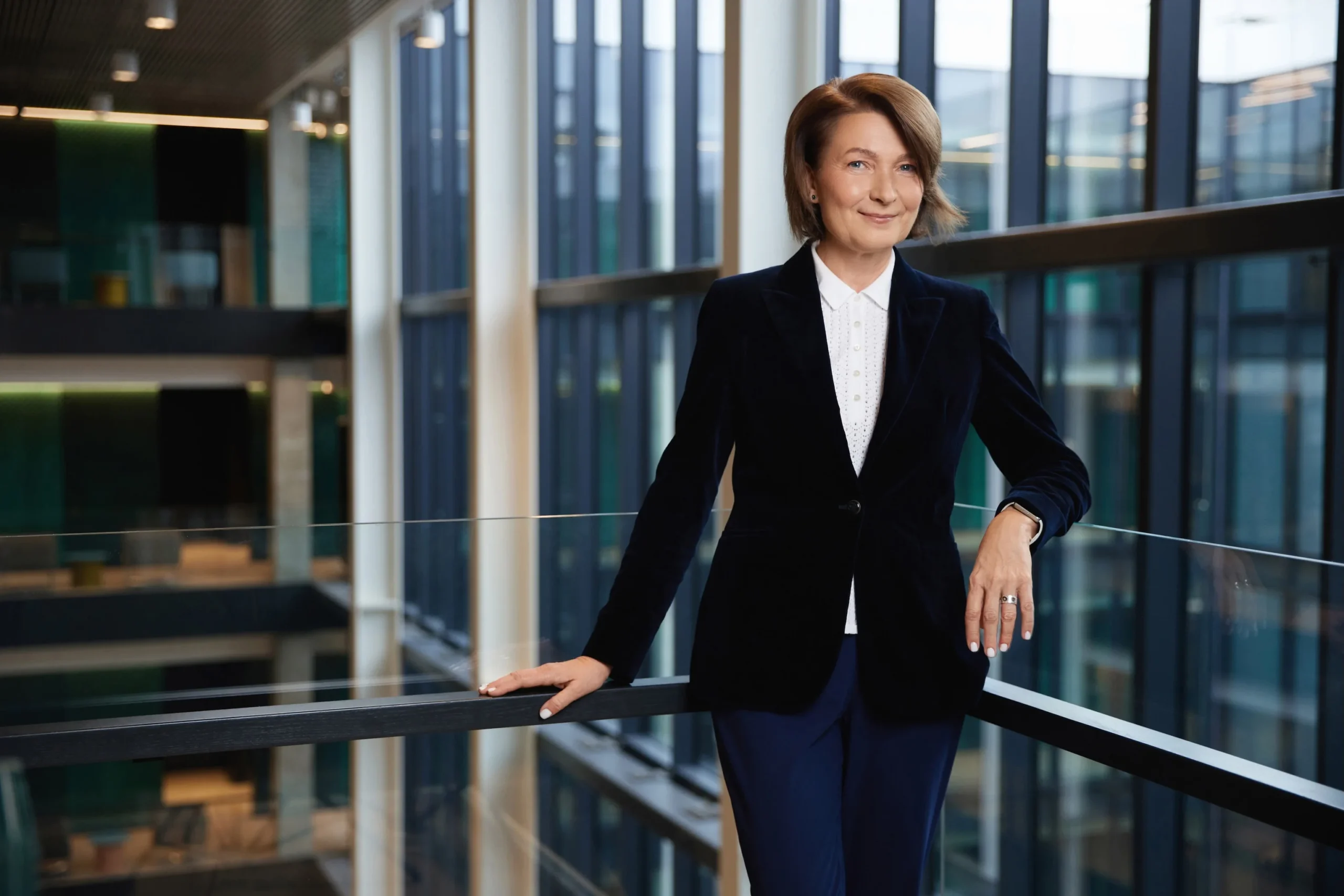 Woman with short brown hair in a dark velvet blazer standing in a modern glass atrium.