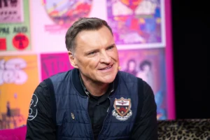 A smiling man wearing a navy blue quilted vest sits in front of colorful posters.