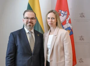 Man in a suit and woman in a beige blazer standing before two Lithuanian flags.